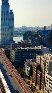 Elevated train line beside residential buildings, Williamsburg, New York. Drone view of red steel viaduct, rooftop terraces and river in soft morning light. Vertical video.