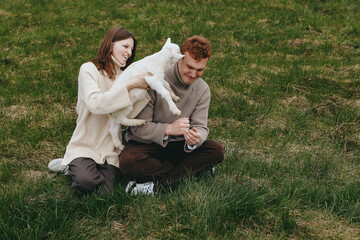 Naklejka premium Cheerful couple outdoors, a white goat kid leaping toward them, gentle interaction, relaxed posture, sunlit grass, natural colors, candid moment, warm mood, rustic setting