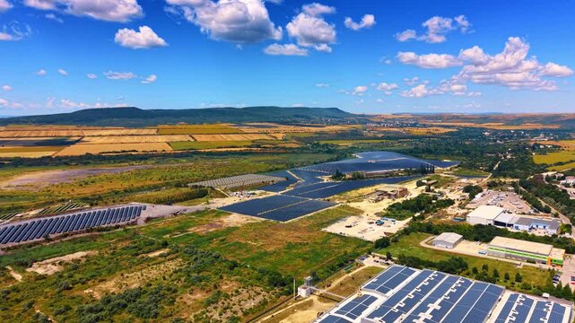 Solar panels cover land in a rural area. Solar panels line the ground in a rural area with fields and mountains nearby. The sky shows some clouds.