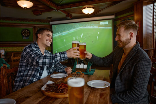 Two white men clinking pints pub, colleagues celebrating goal while watching soccer on large screen