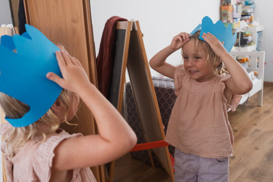  blonde girl wearing a handmade blue paper crown looking at her reflection in a mirror 