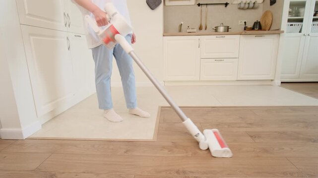 Woman cleaning floor with modern cordless vacuum cleaner in bright kitchen. Housework task and routine maintenance of home. Household appliance for convenient domestic chore and lifestyle.