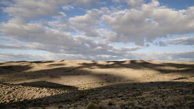Expansive high desert landscape with rolling sand dunes, dramatic cloud shadows and open blue sky.