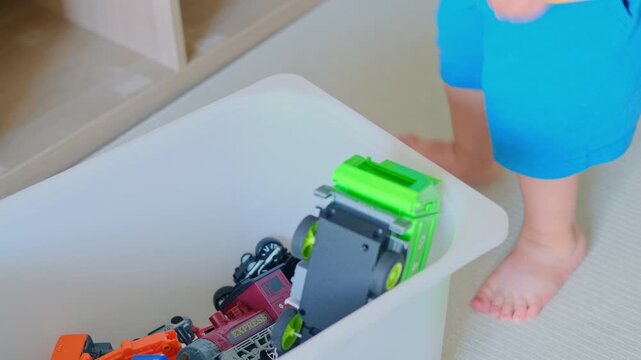 Little child putting green toy truck inside plastic storage box. Kid tidying up bedroom by organizing plastic playthings. Childhood activity and habit of cleaning up room after playing.