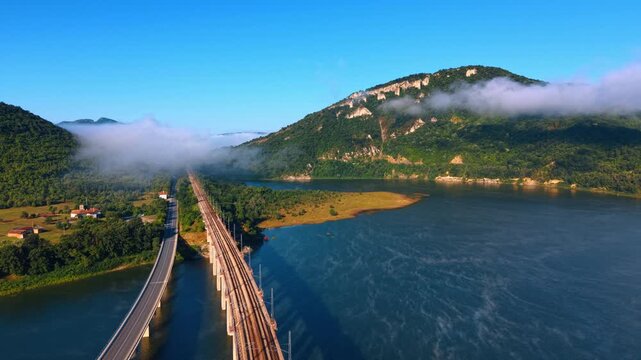 High angle aerial perspective of railway tracks leading across blue water. A parallel two lane road runs alongside concrete train trestles toward misty forested mountains under clear sky.