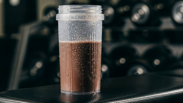 Chocolate protein shake in clear plastic bottle with water droplets on surface sits on black gym bench with blurred dumbbells in background for fitness nutrition.