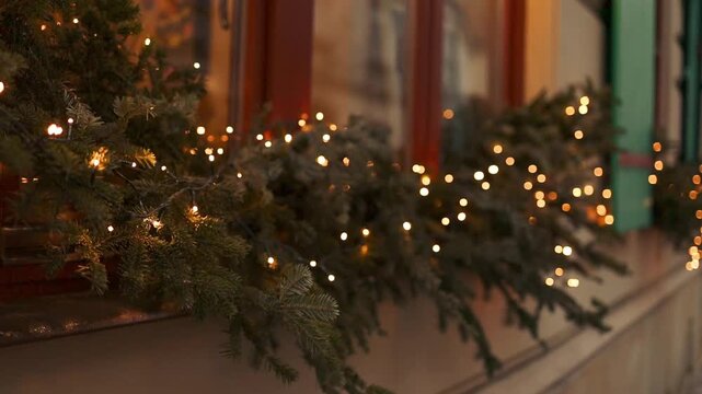 Close up of an outdoor window sill beautifully adorned with lush green pine branches and warm shimmering fairy lights during winter holiday season.