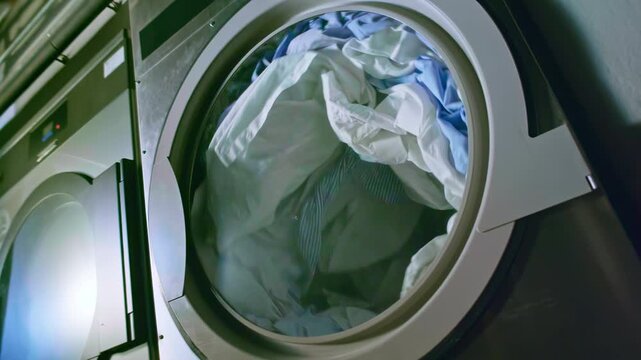 Industrial dryer with freshly washed clothes slowly tumbling inside the rotating drum, smooth slow-motion movement, cinematic look, soft lighting, realistic details, professional atmosphere.
