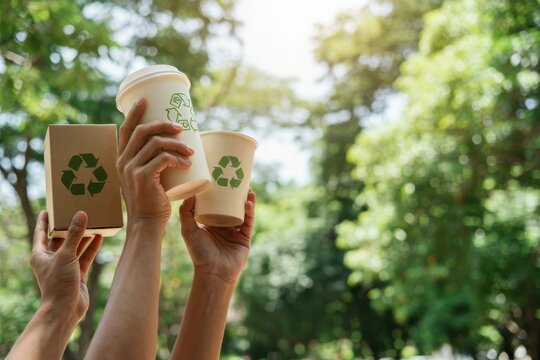 Hands lifting recyclable cups and box against blurry green tree background