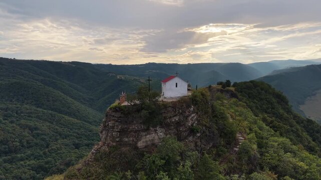 Slow counterclockwise drone orbit around a lonely Orthodox chapel on a rocky outcrop, surrounded by lush green mountains as warm sunset rays break through dramatic clouds
