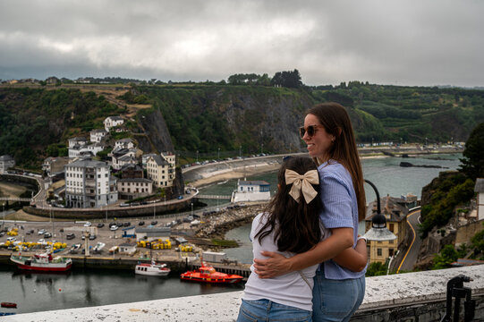 Mother and daughter embracing at viewpoint overlooking scenic coastal fishing town and harbor of Luarca in Asturias, Spain. 