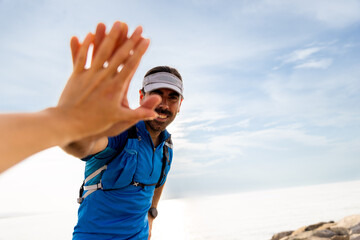 Man in blue shirt and headband smiles. His hand reaches for a high-five. Another person's hand touches his palm. Bright sky and rocks form a backdrop. Ideal for travel or friendship themes © VICTOR