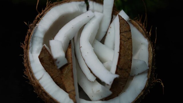 Sliced coconut pieces inside a half coconut shell on a dark background, showing fresh white flesh and brown husk