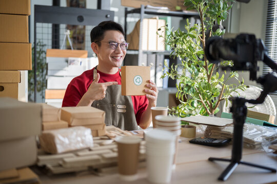 Smiling Asian entrepreneur pointing at a recycled cardboard box, recording a video with a digital camera to promote his sustainable products online.
