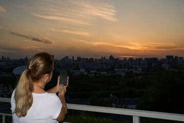 Fototapeta premium Young Woman Using Smartphone To Capture Sunset Skyline From Balcony With City Horizon At Evening.