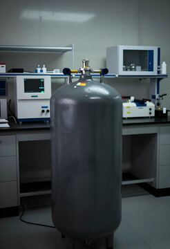 Large gray cryogenic liquid nitrogen dewar tank in a modern laboratory setting with scientific equipment, Various scientific instruments and equipment are visible on the countertops and shelves