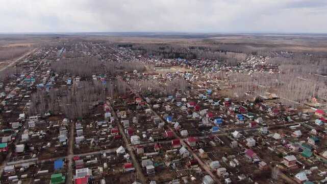 Aerial drone footage flying over a sprawling rural settlement or dacha community. The view reveals hundreds of small country houses with colorful roofs nestled among bare birch forests. A small pond i