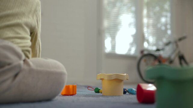 Low-angle home interior showing a young child seated on the left foreground playing with scattered colorful plastic toys on a carpeted floor. Rear view