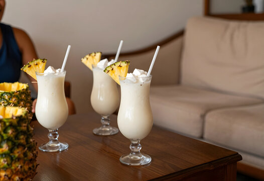 Three pina colada cocktails with pineapple garnish and whipped cream on a wooden table in a relaxed setting, White straws are inserted into the creamy, light-colored drinks