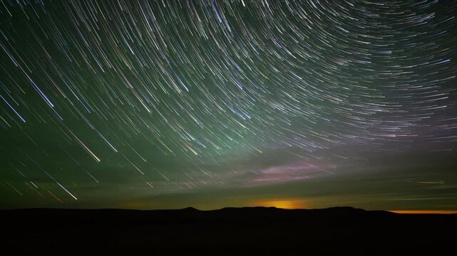 Timelapse of colorful stars with long comet like tales rotating though the night sky with silhouetted hills along the horizon.  The sky starts with sunset colors and changes to weak green Aurora.
