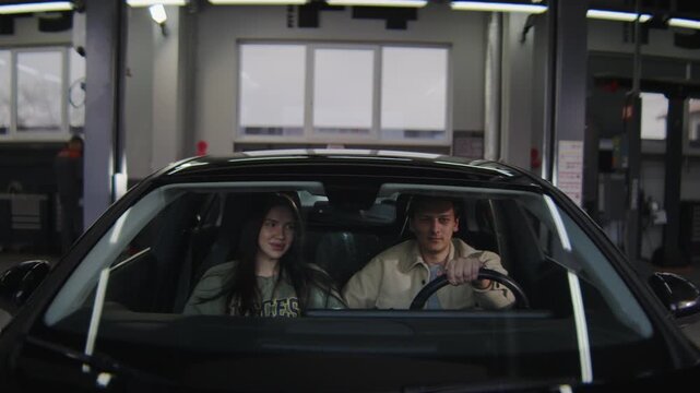 A young, attractive couple is driving away in their new car after buying it. A camera is mounted on the hood of the car, facing the driver and passenger.