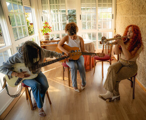Three young women performing together indoors with guitar, bass, and trumpet, seated in bright sunlight beside large windows, creating warm homemade music scene with relaxed artistic energy © carlos