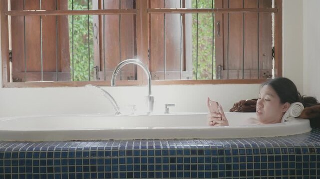 A serene bathroom scene featuring a person enjoying a bath while using a mobile phone. Natural light flows through the window, creating a calming atmosphere. Perfect for illustrating relaxation and mo