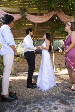 Diverse wedding couple standing under wooden pergola holding hands, displaying white lace gown