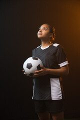 Fototapeta premium African American female soccer player holding black-white ball in black kit, studio-lit, gazing up