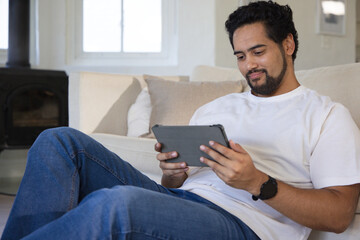 Fototapeta premium African man 30s reclining on light sofa in living room holding gray tablet wearing white tee