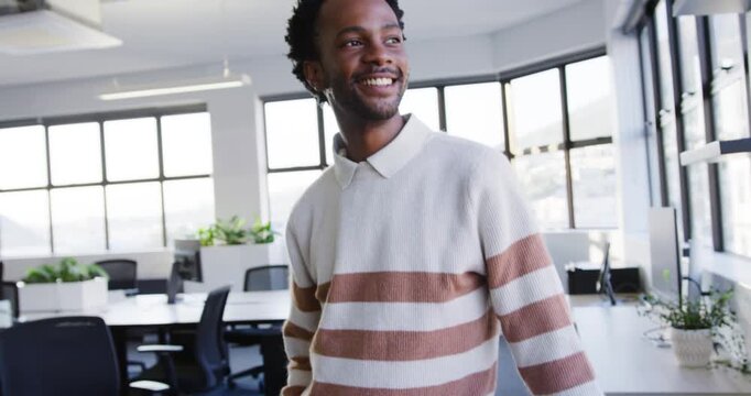 African American man leaning on desk, camera pulling, rising, smiling at windows, in cream sweater