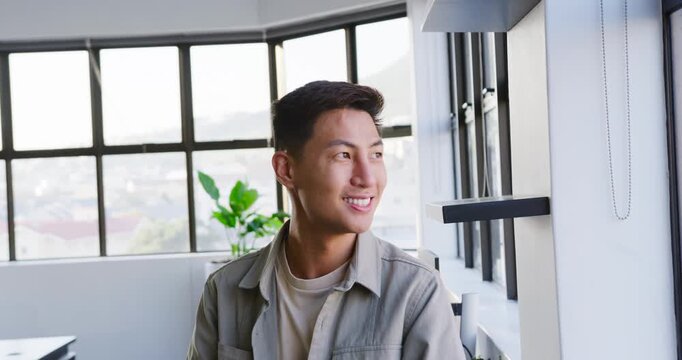 Mid-adult Asian man turning from office windows toward camera, reacting to off-camera cue, smiling