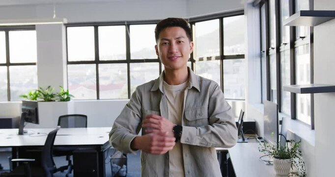 Asian man at office noticing camera pull, rolling sleeve, revealing watch, adopting confident smile