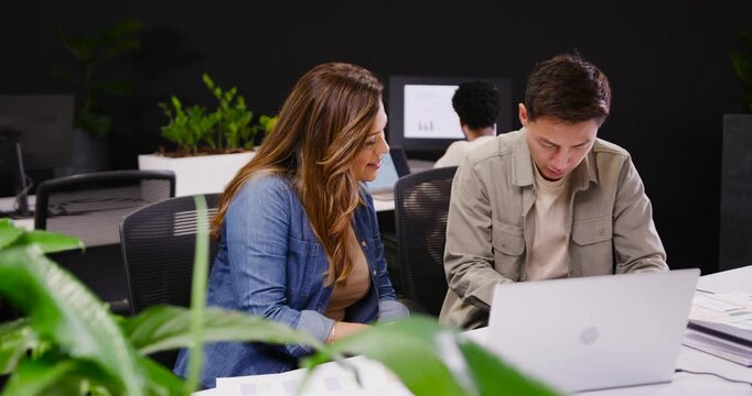 African American woman and Asian man coworkers leaning toward white laptop at desk, collaborating