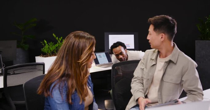Diverse coworkers noticing device, walking to desk, leaning and pointing at tablet to help