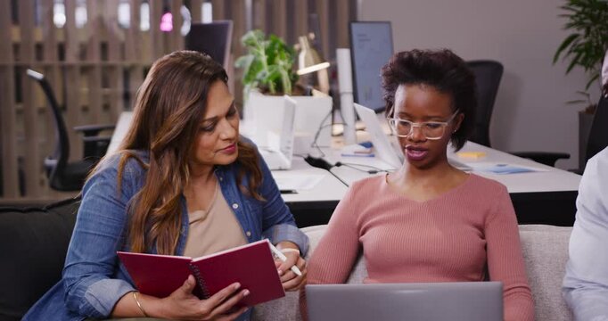Diverse female team reviewing on couch with laptop right starting talk left writing red notebook