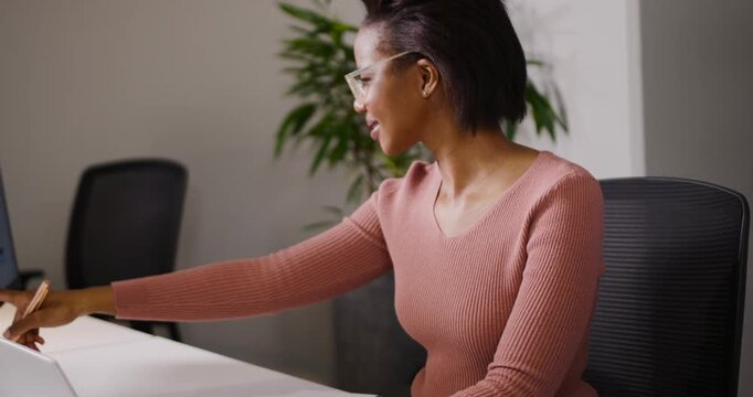 African woman reaching for pen while typing on laptop, writing in notebook, glancing at monitor