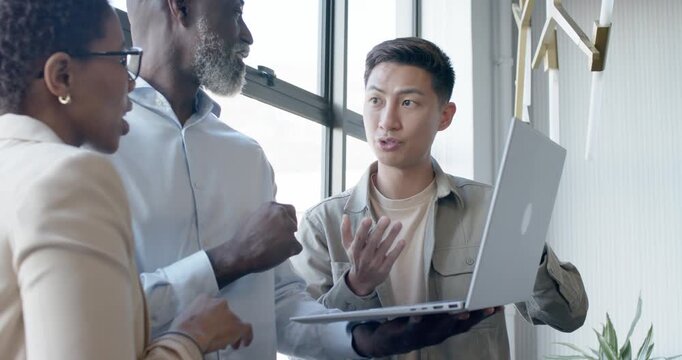 Diverse coworkers with Asian presenter holding silver laptop, leaning and discussing work by window
