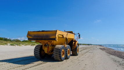 Large yellow dumper truck working on a sandy sea beach during a bright sunny day under blue sky. © Igor