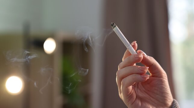 Woman holding burning cigarette at home, closeup. Space for text