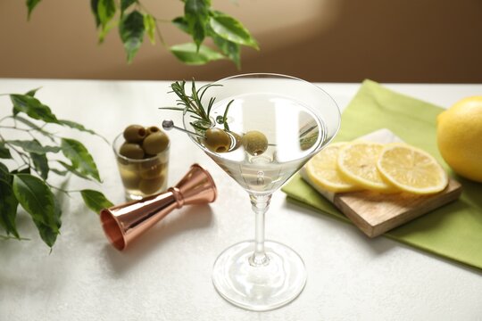 Martini cocktail with olives in glass, lemons, jigger and green plant on white table, closeup