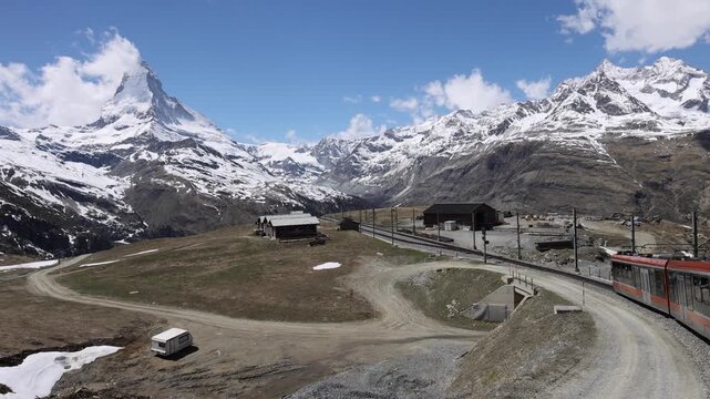 View of the Gornergrat railway train winding through the Swiss Alps with snow-capped peaks and the Matterhorn in the background in Zermatt, Valais, Switzerland.