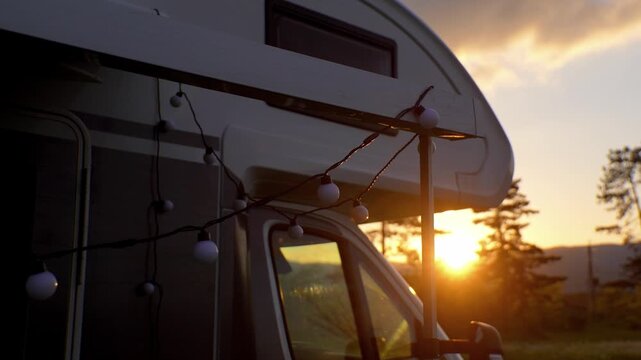 A beautiful golden sun sets behind a parked motorhome, and festive garlands hang on the canopy and create a cozy atmosphere for a perfect trip