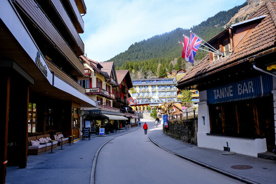 Wengen, Switzerland - March 13, 2026. A ground-level view looking down a paved, pedestrian-only street in the mountain resort village of Wengen, Switzerland. The scene captures local storefronts inclu