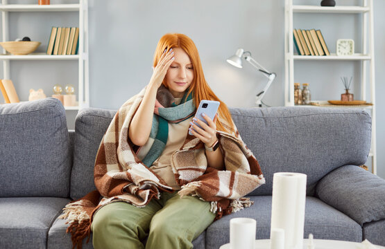 Calling doctor, sad sick young millennial red-haired girl on couch feels fever catching cold, bad flu, influenza, holding phone to make call to therapist, reading internet medical health advice 