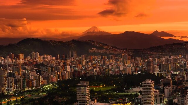 Aerial drone view of Quito, Ecuador at sunset featuring La Carolina Park, illuminated city streets, and Cotopaxi volcano in the distance beneath a dramatic orange and magenta sky.