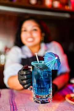 Hispanic woman bartender serving blue cocktail with umbrella