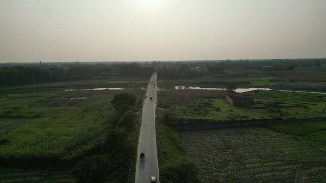 Aerial view of rural road and bridge with three wheeler traffic passing through green fields, drone footage