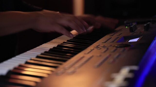 Manos de m&uacute;sico tocando teclado sintetizador en estudio con iluminaci&oacute;n anaranjada y azul.