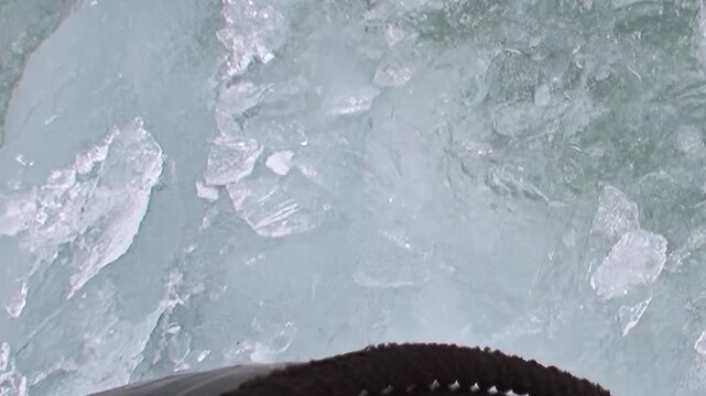 View from a ship navigating the frozen expanses of the Arctic Ocean. The vessel's hull pushes through thick ice and smaller floes, carving a shimmering path through the chilling, turquoise waters.